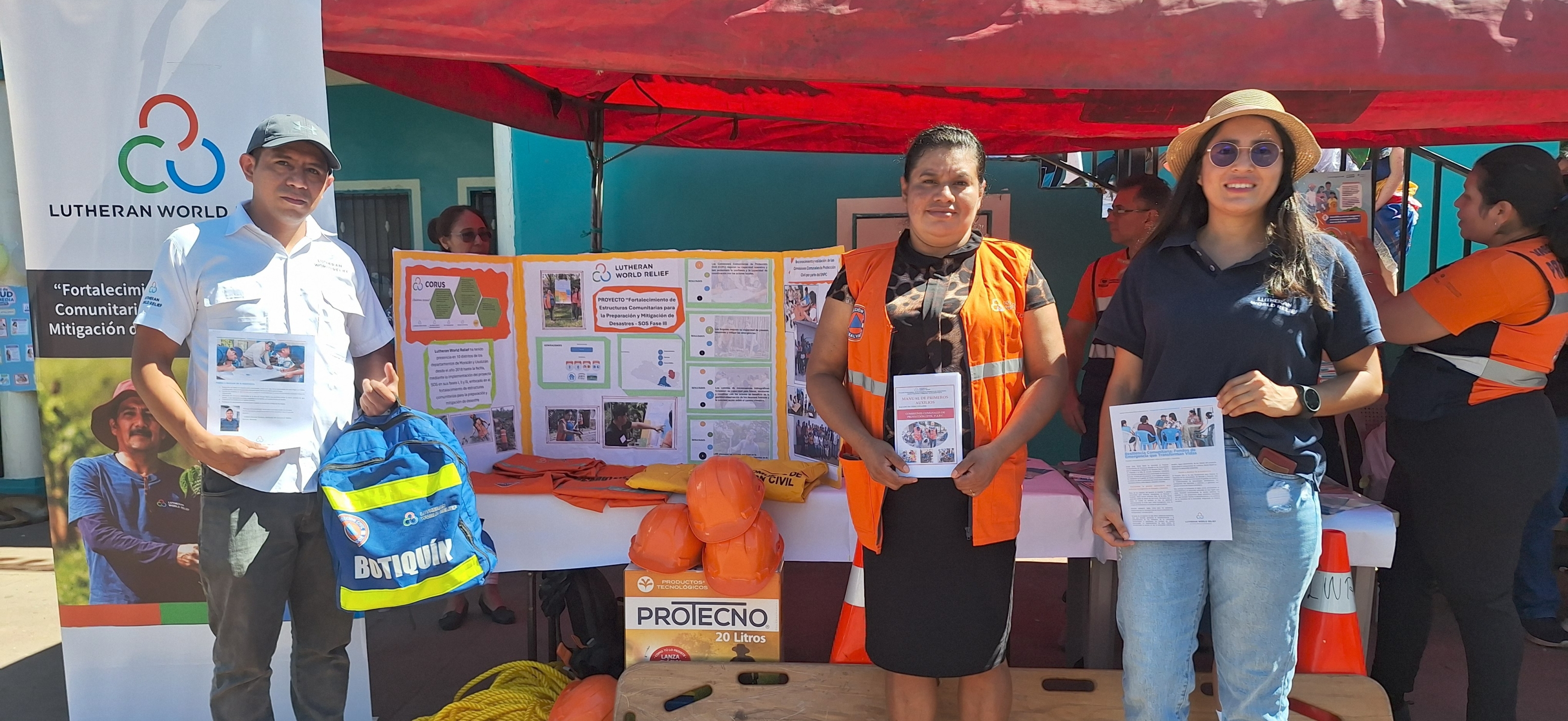 3 people stand in front of an outdoor booth holding up safety gear and guides