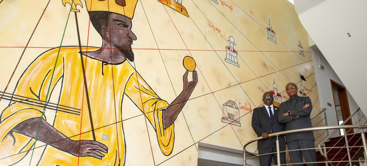 Mahmoud Bah and El-Hassana Kaba stand on stairwell in MANSA BANK next to iconic mural.