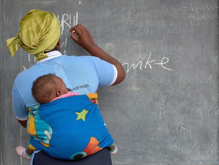 GBV Survivor writes on blackboard carrying her baby wrapped in a blanket on her back