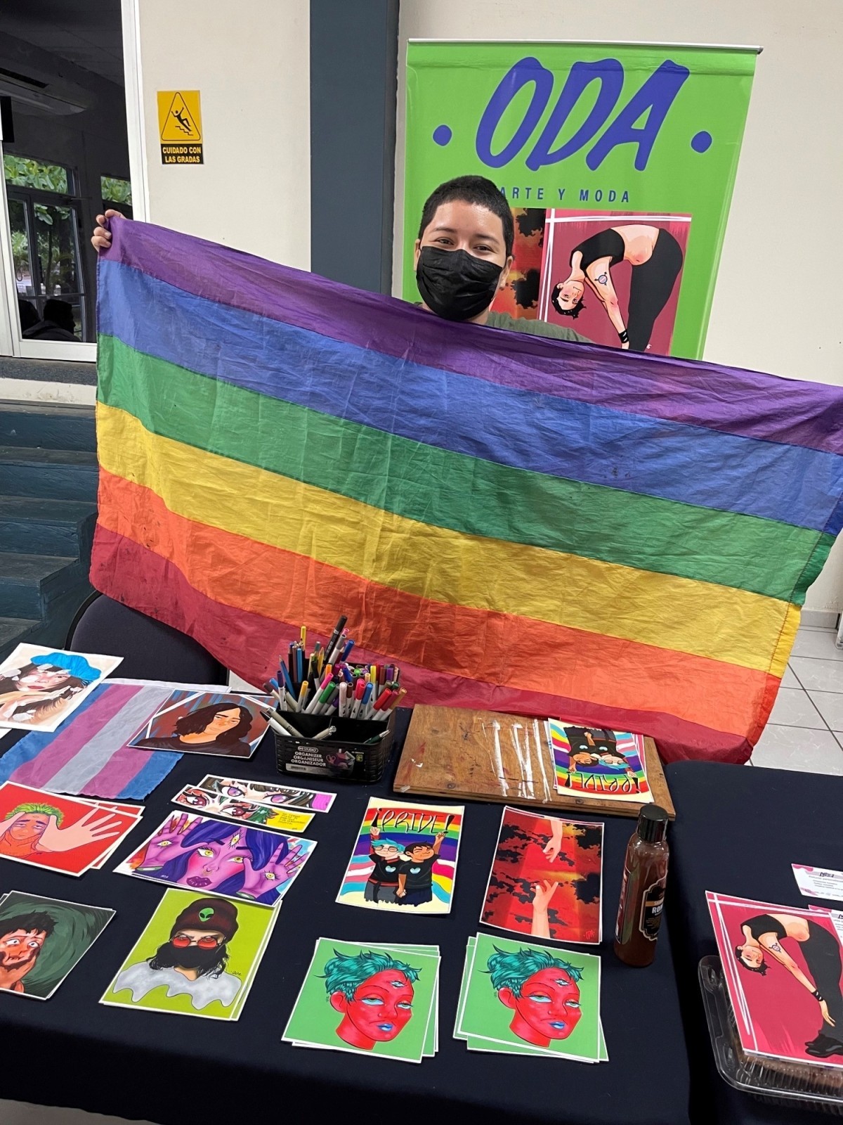 A young man stands behind a table that displays colorful artwork and holds up a rainbow flag