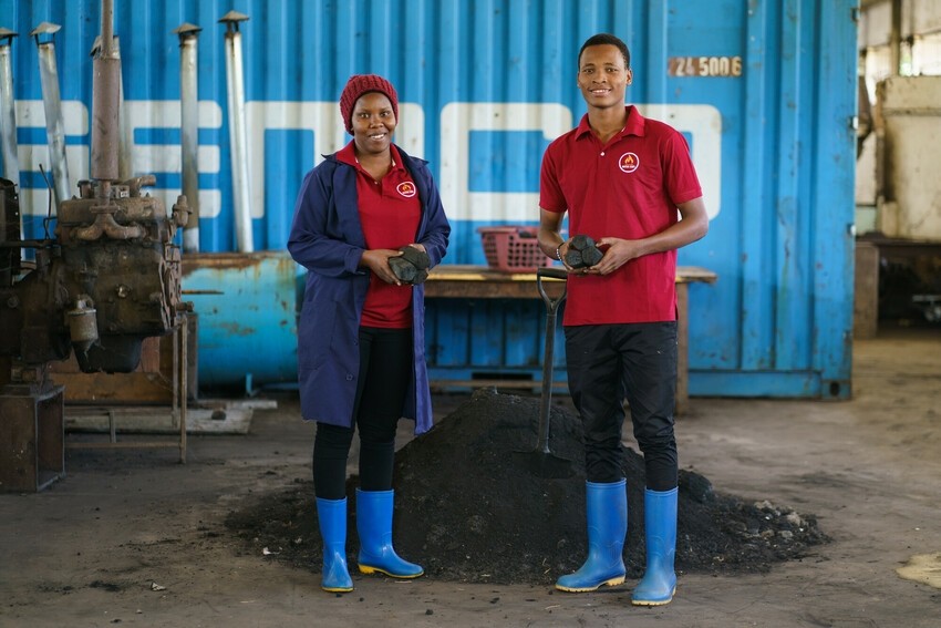 A woman and a man stand in front of a pile of charcoal holding chunks of charcoal