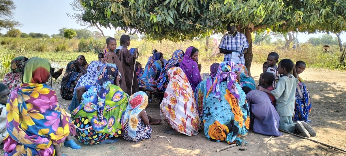 A group of women and children sit outside under a tree listening to a training