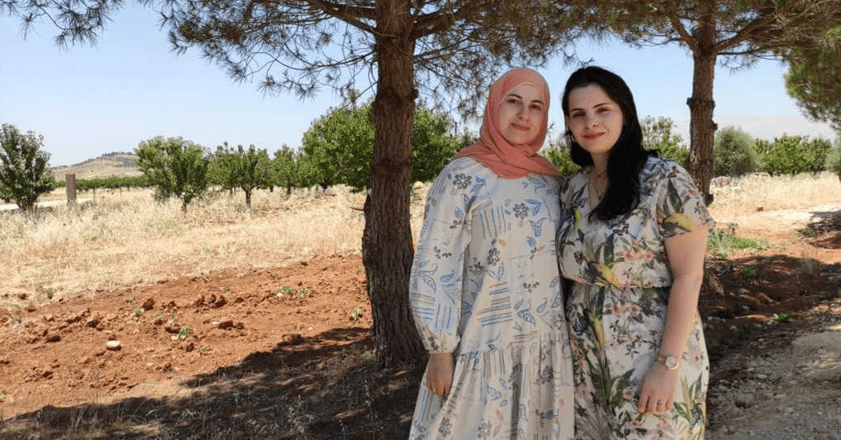 Two sisters stand next to each other smiling outside with trees behind them