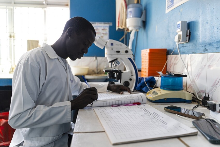A lab technician sits in front of a microscope and notebook