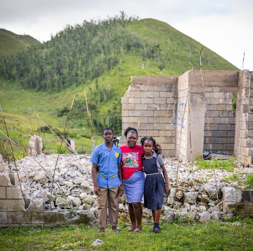 A mom with her 2 children stand in front of a collapsed home