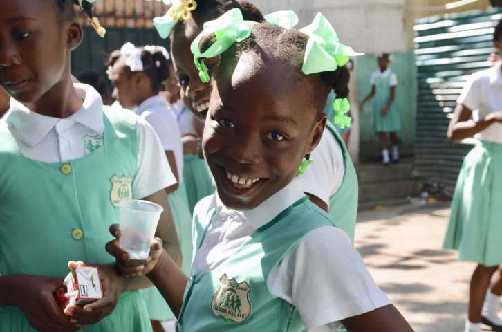 A Haitian girl wearing a mint green and white school uniform smiles while holding a plastic cup.