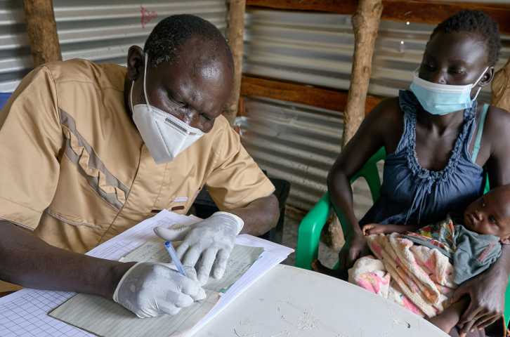 A South Sudanese lab technician writes down health information about an infant who is being held by his mother at a health clinic in South Sudan