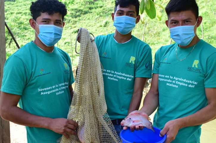 3 Honduran youth stand in front of a pond holding a fishing net and showing off a caught fish