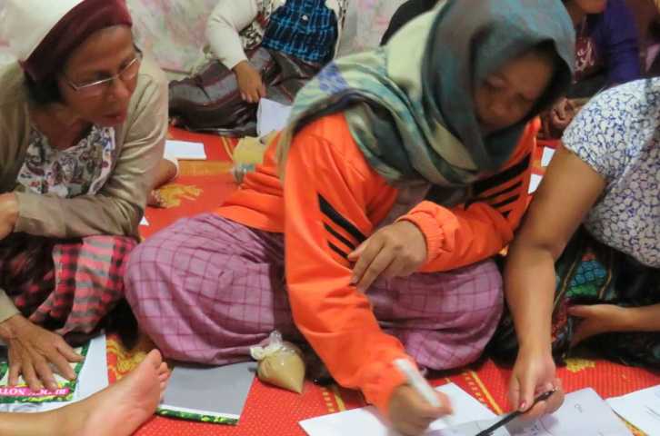 A group of women sit on a bright orange blanket writing on a large sheet of paper together