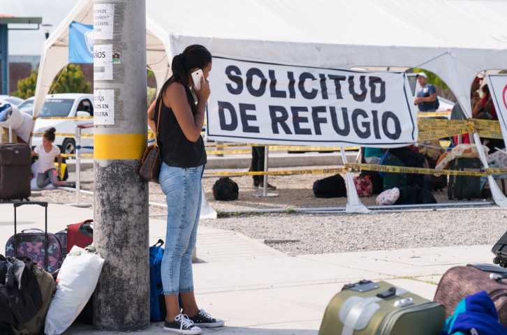 Una mujer permanece junto a una farola hablando por el móvil rodeada de equipajes y pertenencias de compañeros migrantes en el paso fronterizo