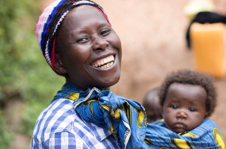A woman with a big smile in a blue and white checkered shirt with her baby tied around her front in a blue and yellow textile