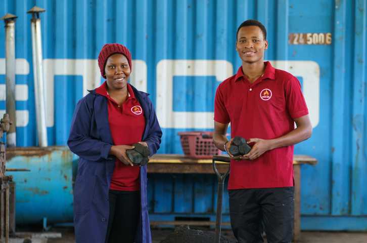 Two young Tanzanian entrepreneurs stand smiling in their workshop while holding charcoal